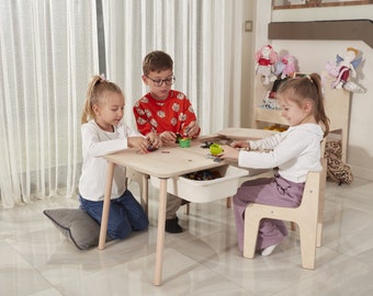 May include: Three children playing with colorful building blocks at a wooden table with a drawer. The table has a light wood finish and the chairs are also made of wood. The children are sitting at the table and are engaged in play.