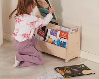 May include: A child is kneeling and reaching for a book from a wooden bookshelf with a handle. The bookshelf is filled with colorful books. The book the child is reaching for has the title "Sayfan Rekolar Kitabi" on the cover.