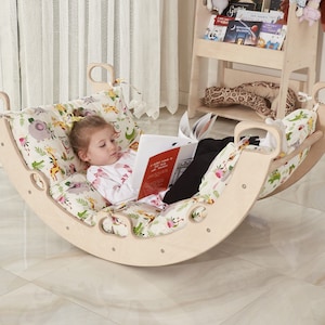 May include: A child sits in a wooden rocking chair with a colorful animal print cushion. The chair is shaped like a crescent moon and has cutouts around the edges. The child is reading a book.