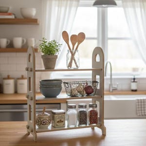 Puede incluir: Una estantería de madera de tres niveles de color blanquecino. Los estantes contienen una variedad de artículos de cocina, incluyendo cuencos, una cesta de alambre con cebollas, frascos de vidrio con especias y una pequeña planta en maceta. Cucharas de madera en un tarro de cristal.