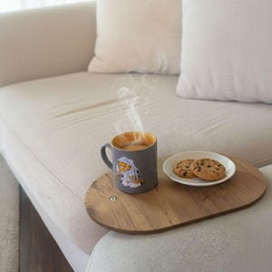 Könnte beinhalten: Eine graue Tasse mit gelbem Innenraum und einem Cartoon-Charakter-Design, gefüllt mit einem heißen Getränk, steht auf einem Holztablett mit einem kleinen Teller mit Schokoladenkeksen. Dampf steigt aus der Tasse auf.