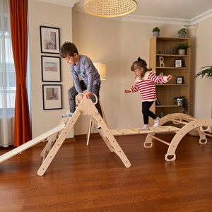 Peut inclure: Deux enfants jouent sur un jeu d'escalade en bois avec un toboggan, un mur d'escalade et un pont. Le jeu d'escalade est en bois naturel et a une finition brun clair. Les enfants s'amusent à jouer sur le jeu d'escalade.