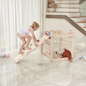 Op de afbeelding: Een houten klimplank met een glijbaan en een ladder. Een kind klimt de glijbaan op. De constructie is gemaakt van natuurlijk hout en heeft een lichtbruine kleur.