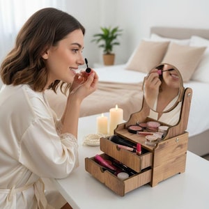 Puede incluir: Un organizador de maquillaje de madera con espejo incorporado. El organizador tiene dos cajones y una balda superior llena de productos de maquillaje. El espejo es redondo y está enmarcado por la estructura de madera. El organizador está sobre una mesa blanca.