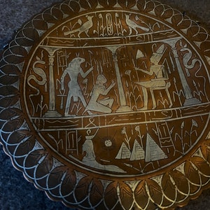 Könnte beinhalten: Ein rundes Kupfertablett mit einem komplizierten Design aus ägyptischen Hieroglyphen und Figuren. Das Tablett ist mit silbernen Akzenten verziert und hat ein zentrales Sonnenmotiv.