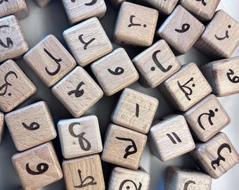 Könnte beinhalten: Eine Sammlung von Holzblöcken, jeder mit einer anderen arabischen Ziffer oder einem Buchstaben, der in Schwarz aufgedruckt ist. Die Blöcke haben eine natürliche Holzfarbe und sind auf einer weißen Oberfläche verstreut.