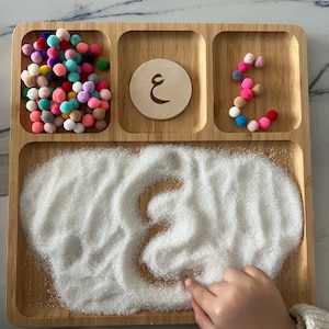 Peut inclure: Un plateau en bois avec quatre compartiments. Le compartiment supérieur gauche contient des pompons colorés. Le compartiment supérieur droit a un cercle en bois avec la lettre arabe 'ع' dessus. Le compartiment inférieur droit contient des pompons colorés disposés en forme de la lettre 'ع'. Le compartiment inférieur gauche contient du sable blanc avec la lettre 'ع' tracée dedans.