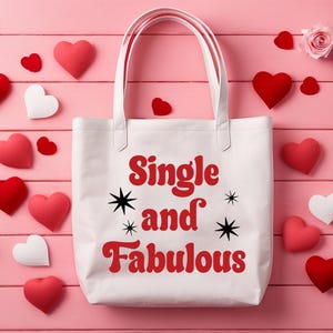 May include: White tote bag with the text "Single and Fabulous" in red, with black starburst accents. The bag is surrounded by red and white heart-shaped decorations and a pink rose on a pink wooden surface.