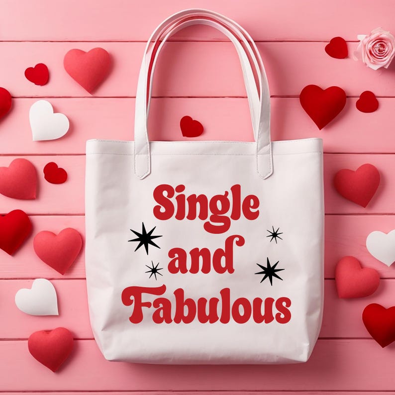 May include: White tote bag with the text "Single and Fabulous" in red, with black starburst accents. The bag is surrounded by red and white heart-shaped decorations and a pink rose on a pink wooden surface.