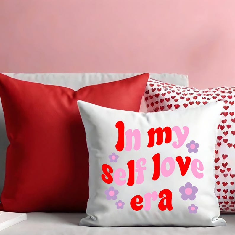 May include: White decorative pillow with the text "In my self love era" in red and pink, with purple flower accents. A solid red pillow is behind it, along with a pillow with a heart pattern.
