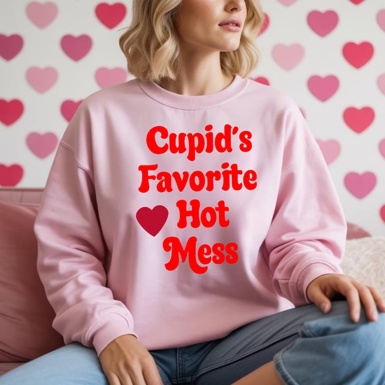 May include: A light pink sweatshirt with the text "Cupid's Favorite Hot Mess" in red. The design includes a red heart. The background features a pattern of pink and red hearts.