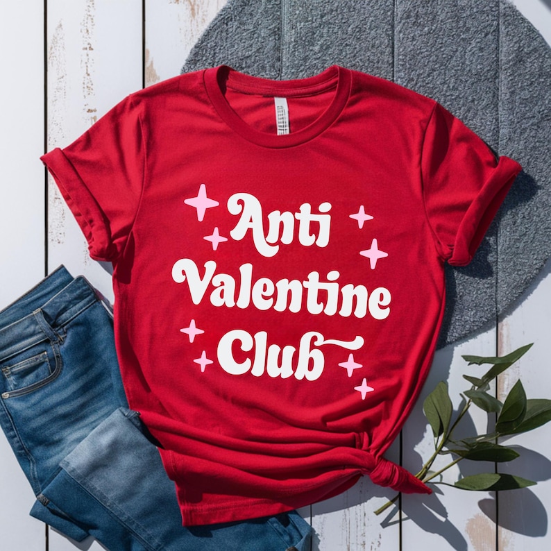 May include: A red t-shirt with the text "Anti Valentine Club" in white, surrounded by pink starbursts. The shirt is styled with blue jeans and a sprig of greenery.