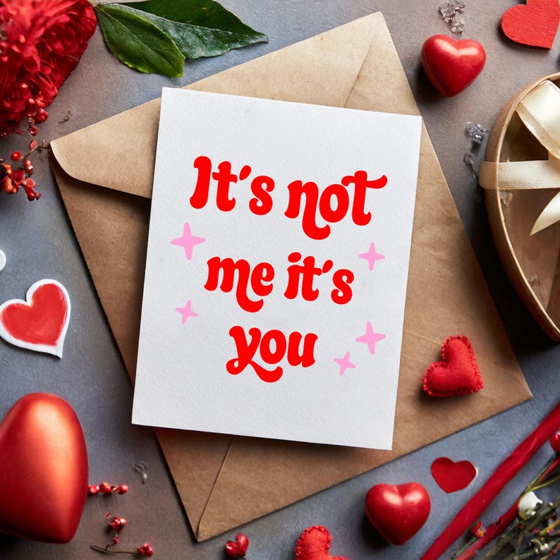 May include: A white greeting card with the text "It's not me it's you" in red, with pink star accents, rests on a brown envelope. The image includes red heart-shaped decorations and a gift box, suggesting a Valentine's Day theme.