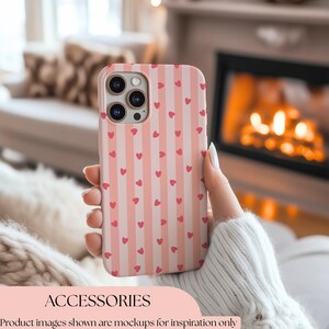 May include: A pink and white striped phone case with small red hearts. The case is held in a hand wearing a white jumper. The word "ACCESSORIES" is at the bottom of the image.