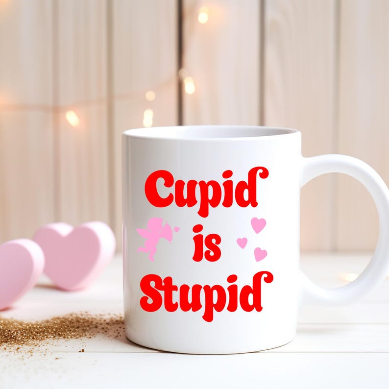 May include: White ceramic mug with the text "Cupid is Stupid" in red, along with a pink cupid and hearts. The mug has a curved handle and is set against a blurred background with decorative lights and pink heart-shaped objects.