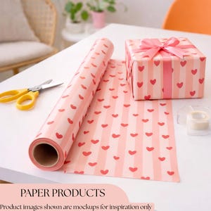 May include: A roll of pink wrapping paper with red hearts and vertical stripes, alongside a wrapped gift box with a pink bow. Yellow scissors and tape are on the white surface. The text "PAPER PRODUCTS" is at the bottom.