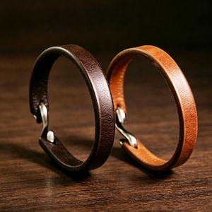 May include: Two circular bracelets made of leather, one in a dark brown shade and the other in a lighter tan colour. Each bracelet features a smooth, rounded band design with a metallic silver-toned hook clasp mechanism visible at the closure point.