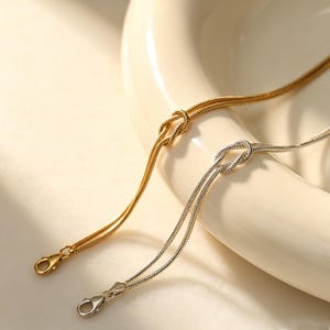 Könnte beinhalten: Zwei zarte Schlangenkettchen-Armbänder, eines in Goldoptik und eines in Silberoptik, jeweils mit einem Knoten-Detail in der Mitte. Beide Armbänder bestehen aus einer glatten, flexiblen Kette und haben einen kleinen Karabinerverschluss am Ende.