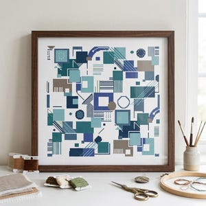 Op de afbeelding: Een ingelijst kruissteek kunstwerk met een geometrische compositie van vierkanten, rechthoeken en cirkels in tinten blauw, groenblauw en beige. Het ontwerp bevat diverse patronen zoals strepen, stippen en rasters op een witte stof.