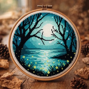 May include: A cross-stitch embroidery hoop featuring a nighttime landscape. The design depicts a full moon over a body of water, framed by silhouetted trees. The color palette includes shades of blue, black, and yellow, with a wooden hoop.
