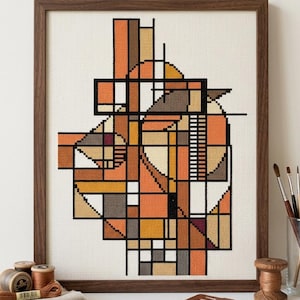 Op de afbeelding: Een ingelijst kruissteek borduurwerk met een abstracte geometrische compositie van elkaar kruisende lijnen en vormen in tinten oranje, bruin, beige en zwart op een crèmekleurige stoffen achtergrond, geplaatst in een donkere houten lijst.