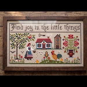 Może przedstawiać: Wyszywana krzyżykami próbka w drewnianej ramie, przedstawiająca sielską scenę z domem, kwiatami i postacią. Tekst "Find joy in the little things" jest wyszyty u góry. Paleta kolorów obejmuje czerwienie, zielenie i brązy.