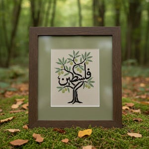 Op de afbeelding: Een ingelijst kruissteekkunstwerk met een gestileerde olijfboom met het Arabische woord "Palestina" in zwart. De boom heeft groene bladeren en olijven. Het kunstwerk staat tegen een lichtgroene achtergrond en is ingelijst.