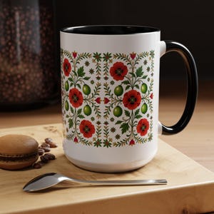 May include: White ceramic mug with a black handle and rim, featuring a floral pattern of red poppies, green berries, and green leaves. The mug is on a wooden surface with a spoon and a macaron.