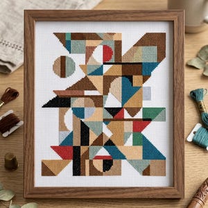 Può includere: Un ricamo a punto croce incorniciato che presenta un motivo geometrico astratto composto da triangoli, cerchi e rettangoli in tonalità marrone, blu, rosso, verde e beige su tela Aida bianca, racchiuso in una cornice di legno.