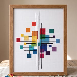 May include: A framed cross-stitch artwork featuring a geometric design of colorful squares and black lines against a white background. The squares are arranged in a spectrum of colors, from red to purple, within a wooden frame. The artwork is displayed on a wooden surface.