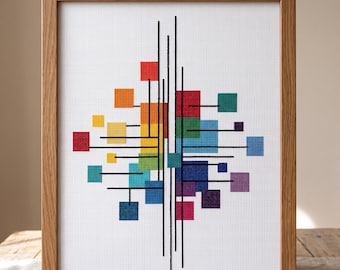 May include: A framed cross-stitch artwork featuring a geometric design of colorful squares and black lines against a white background. The squares are arranged in a spectrum of colors, from red to purple, within a wooden frame. The artwork is displayed on a wooden surface.