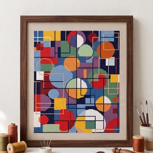 May include: A framed abstract cross-stitch art piece. The artwork features a geometric design with overlapping circles, squares, and rectangles in various colors, including red, blue, orange, green, and yellow. The frame is made of dark brown wood.