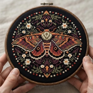Può includere: Un telaio da ricamo circolare in legno contenente un disegno a punto croce su tessuto nero. Il motivo presenta una falena centrale con ali arancioni, rosse e viola, circondata da piccoli fiori bianchi e fogliame verde in modo simmetrico.