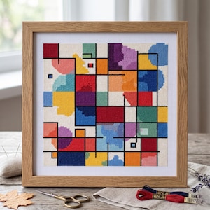 Op de afbeelding: Een vierkant kruissteek borduurwerk met een raster van zwarte lijnen gevuld met diverse geometrische vormen in rood, geel, blauw, groen, paars en oranje, geplaatst in een lichthouten lijst tegen een witte stoffen achtergrond.