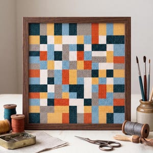 May include: A framed cross-stitch piece featuring a geometric design of multi-coloured squares. The colours include blue, orange, yellow, grey, and white. Crafting supplies such as thread spools, scissors, and paintbrushes are visible on the white surface.