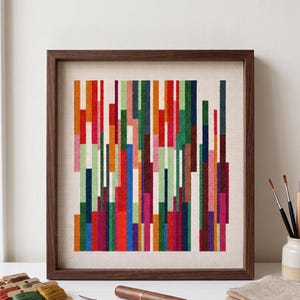 Op de afbeelding: Een ingelijst kruissteekkunstwerk met een abstract ontwerp van verticale balken in levendige kleuren, waaronder rood, oranje, groen en blauw. Het kunstwerk is geplaatst tegen een neutrale achtergrond en ingelijst in een donkerbruine houten lijst. Een moderne, kleurrijke wanddecoratie.