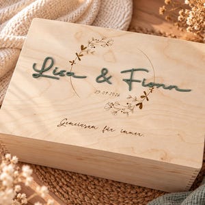 Könnte beinhalten: Eine rechteckige Holzkiste in hellem Farbton mit dem erhabenen grünen Schriftzug Lisa & Fionn, dem Datum 25.09.2026 und dem Text Gemeinsam für immer. Der Deckel ist mit einem feinen, eingravierten Blumenkranz verziert, der die Namen umrahmt.