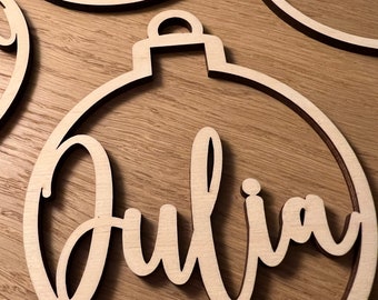 Könnte beinhalten: Drei Holzornamente mit darauf geschriebenen Namen. Die Ornamente haben die Form eines Kreises mit einer kleinen Schlaufe oben. Die Namen sind "Luna", "Don" und "Julia".