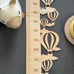 Könnte beinhalten: Ein hölzerner Höhenmesser mit einer in Zentimetern von 70 bis 160 markierten Skala. Der Höhenmesser ist mit einem Design aus Heißluftballons mit Wolken verziert. Der Name "Mara" ist auf dem Höhenmesser geschrieben.