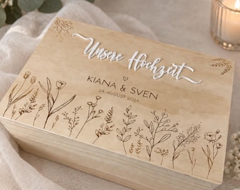 Könnte beinhalten: Eine rechteckige Holzkiste in hellem Farbton mit weißem 3D-Schriftzug "Unsere Hochzeit" über den eingravierten Namen "KIANA & SVEN" und dem Datum "24. AUGUST 2024". Der Deckel ist mit feinen Strichzeichnungen von Wildblumen verziert.