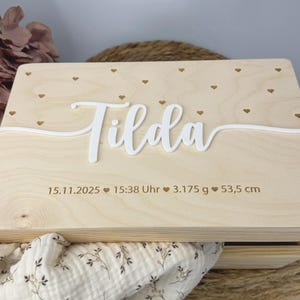 Könnte beinhalten: Eine hellfarbene Holzkiste mit dem Namen "Tilda" in weißer Schrift. Die Kiste ist mit kleinen Herzformen verziert und enthält eingravierte Details: Datum, Uhrzeit, Gewicht (3.175 g) und Länge (53.5 cm).