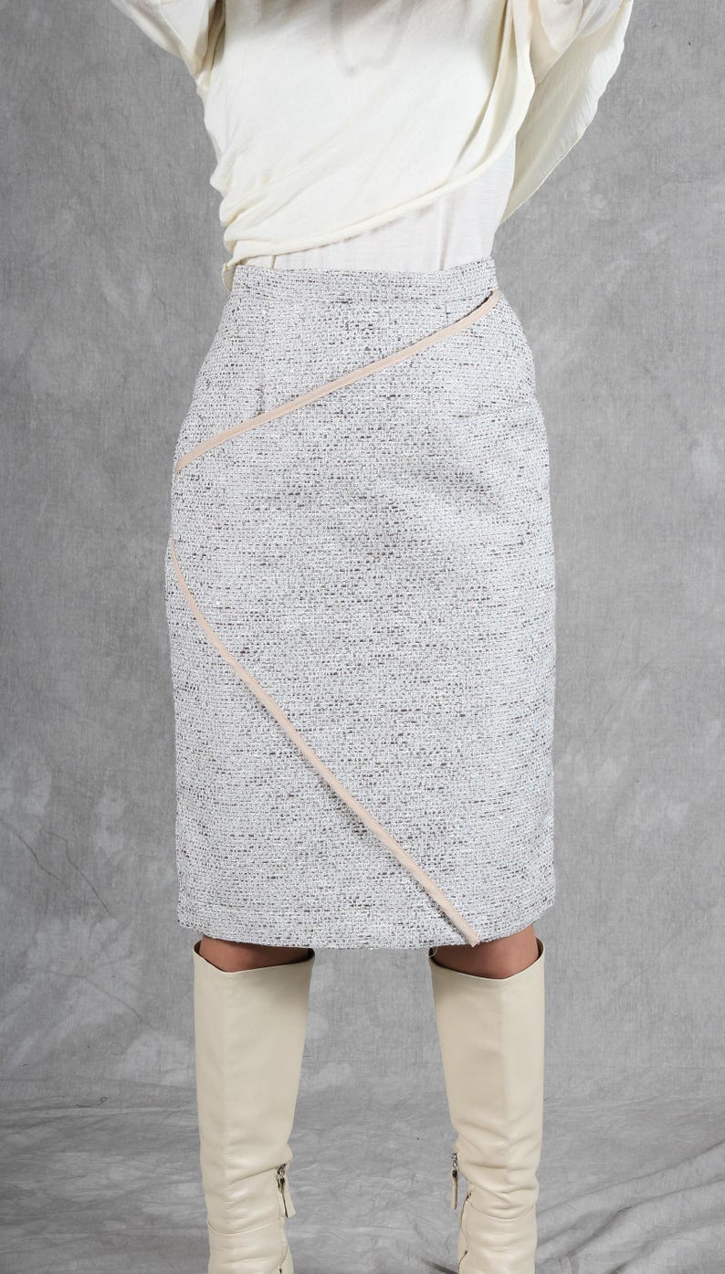 K&ouml;nnte beinhalten: Ein knielanger Bleistiftrock aus strukturiertem, wei&szlig;-schwarzem Tweed-Stoff. Der Rock zeichnet sich durch eine markante diagonale Naht aus, die mit einer glatten, hellbeigen Paspelierung von der Taille bis zum unteren Saum verziert ist.
