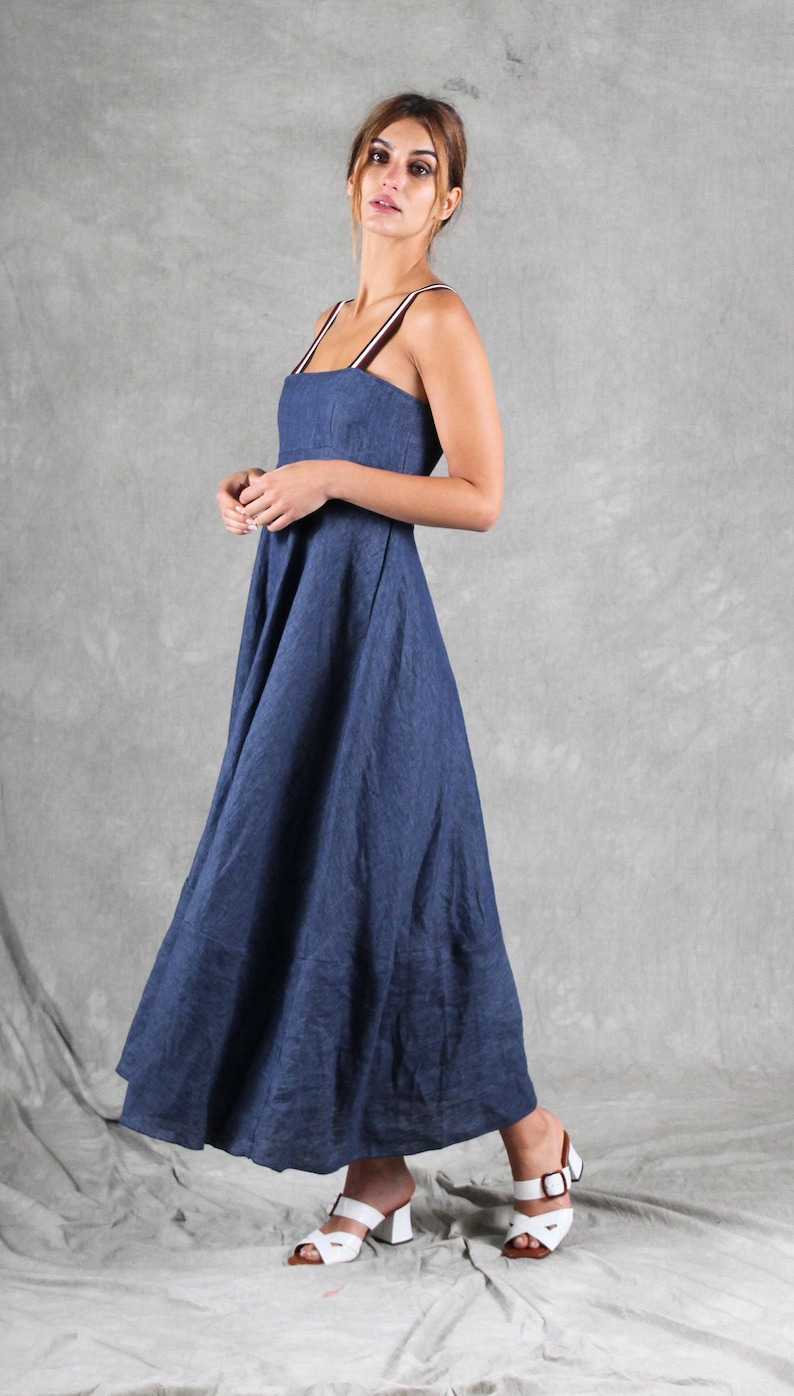 May include: A floor-length navy blue dress with a square neckline and thin, striped shoulder straps in white, brown, and black. The garment features a fitted bodice that transitions into a full, flowing skirt, paired with white block-heeled sandals.