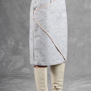 K&ouml;nnte beinhalten: Ein knielanger Rock aus gesprenkeltem grau-wei&szlig;em Tweed-Stoff mit einer diagonalen Naht und hellbeiger Paspelierung. Der Rock hat einen strukturierten Bund und eine gerade Silhouette, kombiniert mit cremefarbenen, kniehohen Lederstiefeln.