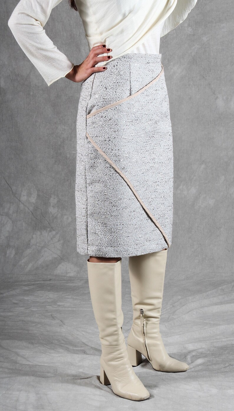 K&ouml;nnte beinhalten: Ein knielanger Rock aus gesprenkeltem grau-wei&szlig;em Tweed-Stoff mit einer diagonalen Naht und hellbeiger Paspelierung. Der Rock hat einen strukturierten Bund und eine gerade Silhouette, kombiniert mit cremefarbenen, kniehohen Lederstiefeln.