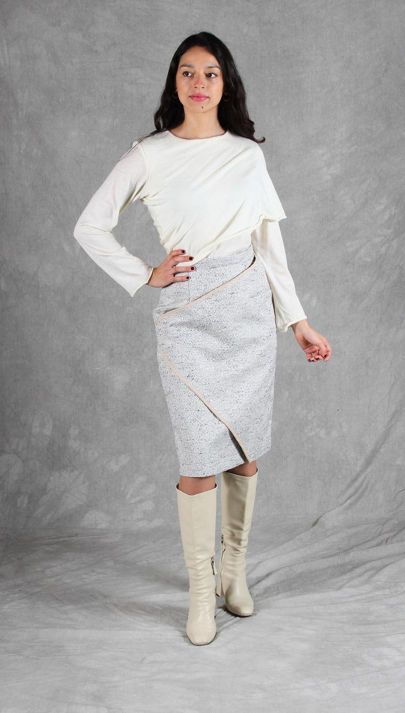 K&ouml;nnte beinhalten: Ein lang&auml;rmeliges cremefarbenes Oberteil mit drapiertem Ausschnitt kombiniert mit einem knielangen grauen Tweedrock, der ein diagonal gewickeltes Vorderteil mit hellbeiger Kante aufweist, sowie cremefarbene kniehohe Lederstiefel.