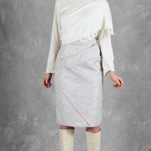 K&ouml;nnte beinhalten: Eine cremefarbene Langarmbluse mit drapiertem Ausschnitt und ein knielanger grauer Tweedrock mit diagonalem hellrosa Zierstreifen, kombiniert mit cremefarbenen kniehohen Stiefeln. Das Outfit besteht aus einem einfarbigen Oberteil und einem strukturierten Rock.