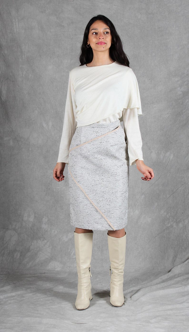 K&ouml;nnte beinhalten: Eine cremefarbene Langarmbluse mit drapiertem Ausschnitt und ein knielanger grauer Tweedrock mit diagonalem hellrosa Zierstreifen, kombiniert mit cremefarbenen kniehohen Stiefeln. Das Outfit besteht aus einem einfarbigen Oberteil und einem strukturierten Rock.