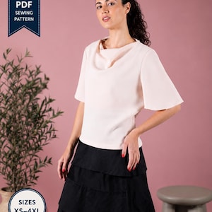 Peut inclure: Un chemisier rose pâle avec un col bénitier et des manches courtes évasées, associé à une jupe noire à étages. L'image comprend une bannière bleu foncé avec le texte PDF SEWING PATTERN et un badge circulaire avec le texte SIZES XS-4XL.