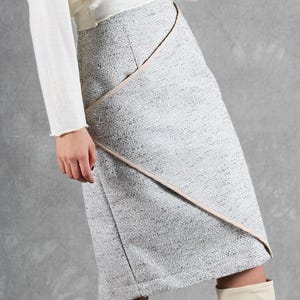 K&ouml;nnte beinhalten: Ein knielanger Rock aus strukturiertem grau-wei&szlig;em Tweed-Stoff. Der Rock verf&uuml;gt &uuml;ber ein Vorderteil in Wickeloptik mit einer diagonalen Naht, die durch eine glatte, hellbeige Borte akzentuiert wird, die von der Taille bis zum Saum verl&auml;uft.